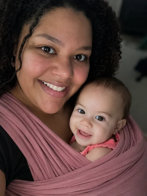 Smiling caregiver holding her baby in a pink wrap carrier during The First Dance babywearing class